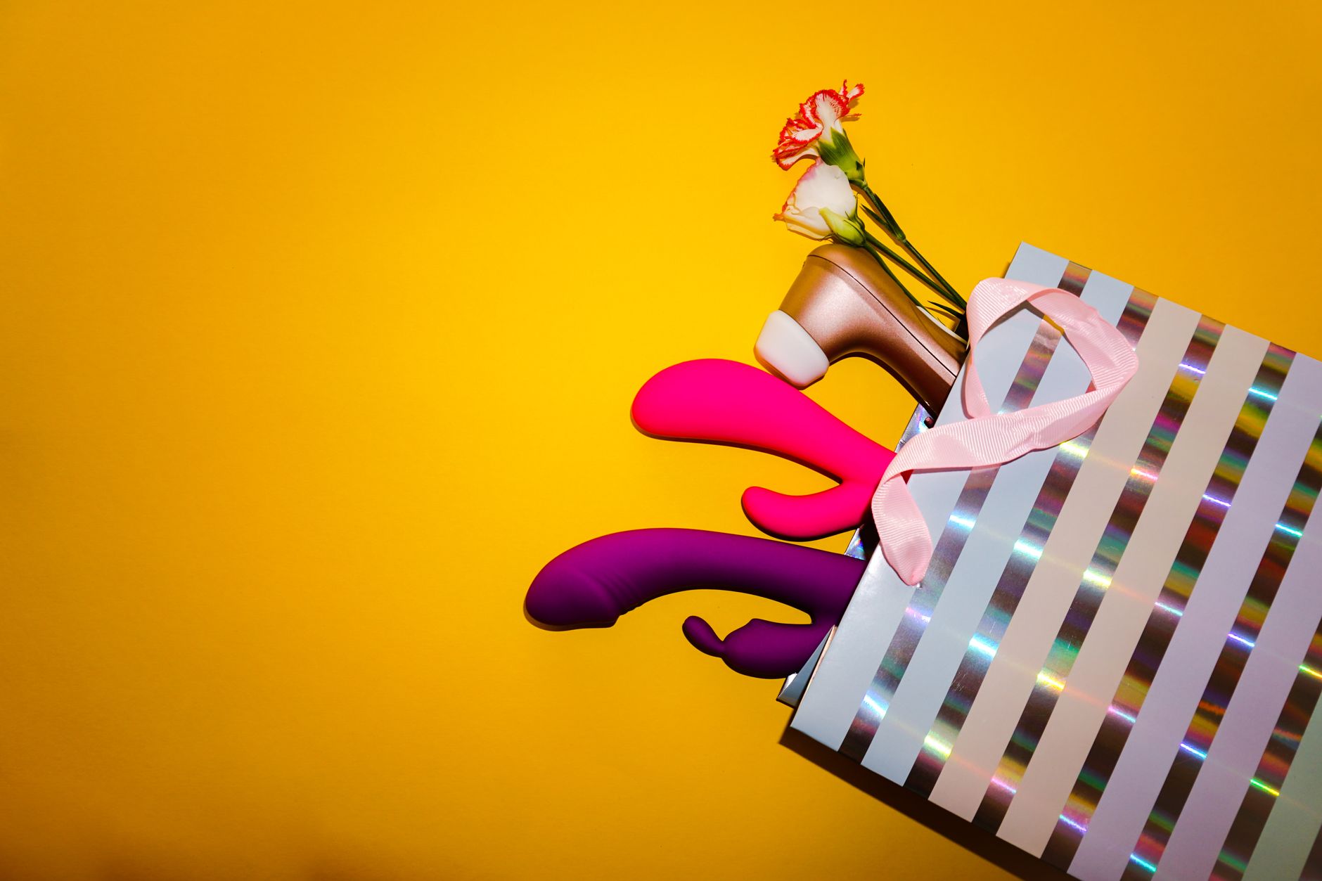 Colorful vibrators and intimate wellness items in a holographic gift bag against a bold yellow background.